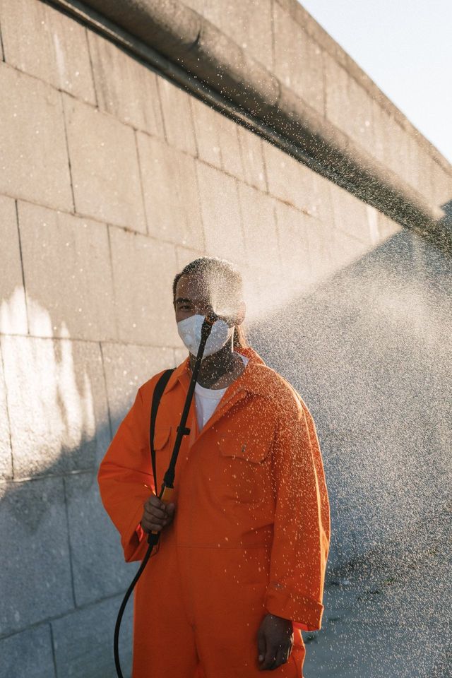 Person in orange jumpsuit and face mask spraying water, standing in front of a stone wall.