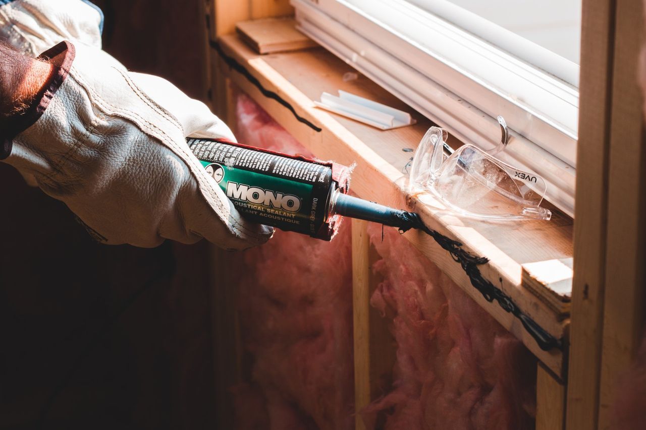 Person caulking around a window frame with a tube of sealant, wearing gloves; construction site.