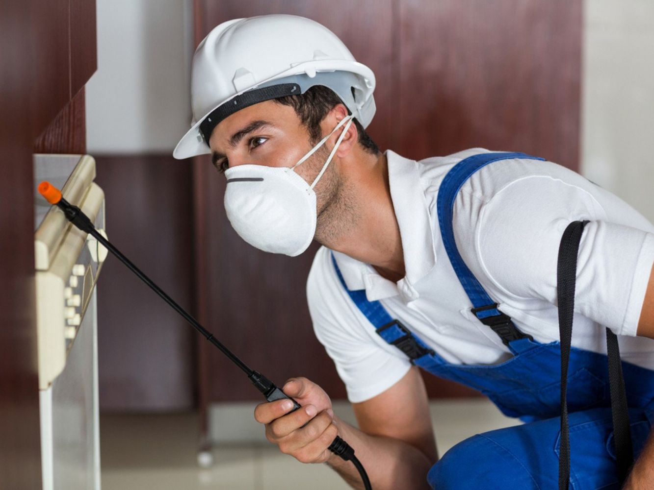 Pest control worker spraying insecticide in a cabinet, wearing a respirator, hard hat, and blue overalls.